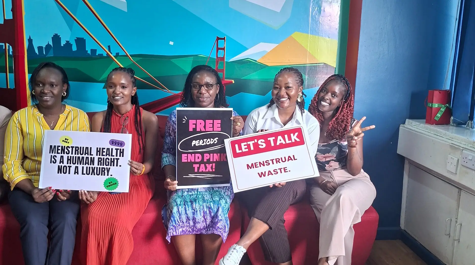 Five women sit together holding menstrual health and dignity placards against a bright mural.