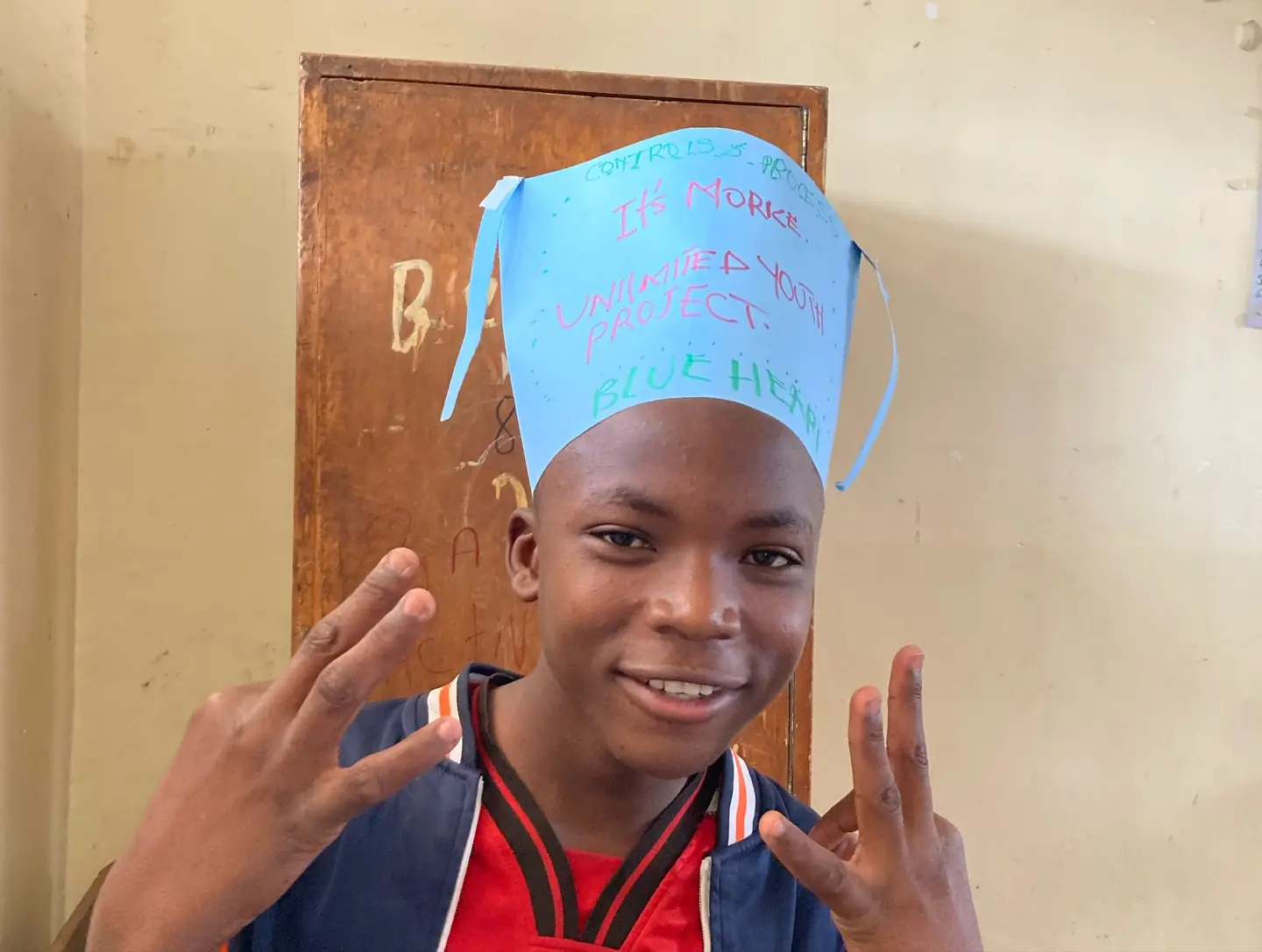 A teenager smiles and makes a peace sign while wearing a handmade blue paper hat.