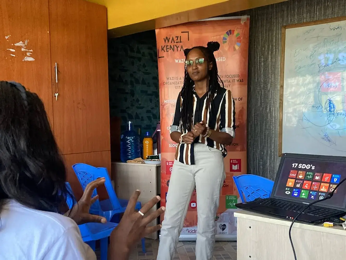 A facilitator stands beside a whiteboard and a laptop displaying the Sustainable Development Goals.