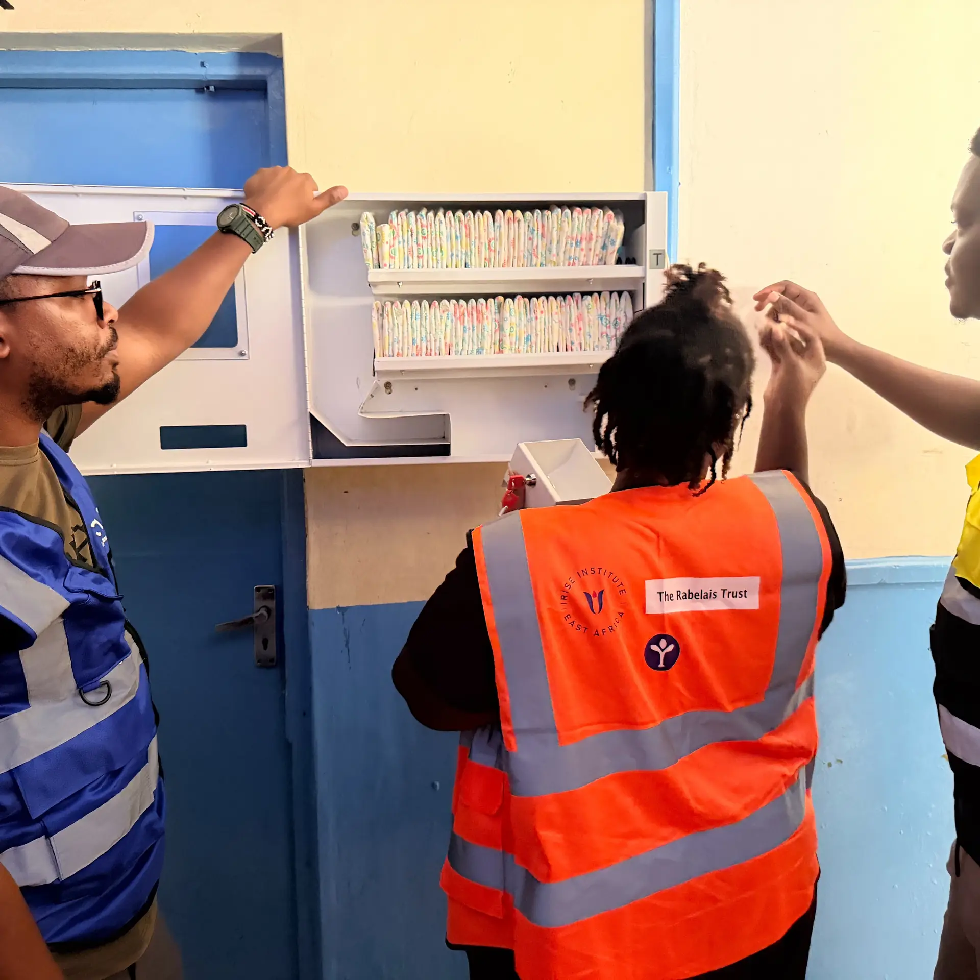 Wazi and CAEL volunteers open a stocked wall dispenser in a blue and yellow corridor.