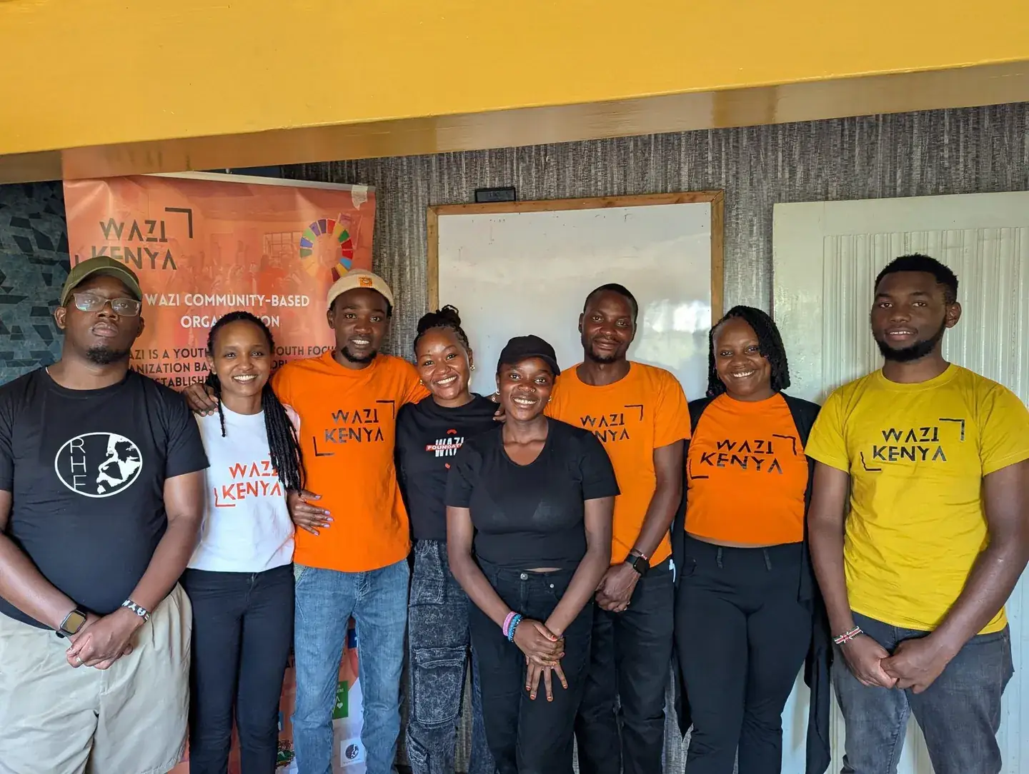 Seven members of the Wazi Kenya team in orange, black and yellow Wazi shirts stand together in front of a Wazi banner.