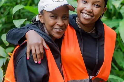Two teammates in orange vests hug and smile among green forest leaves.