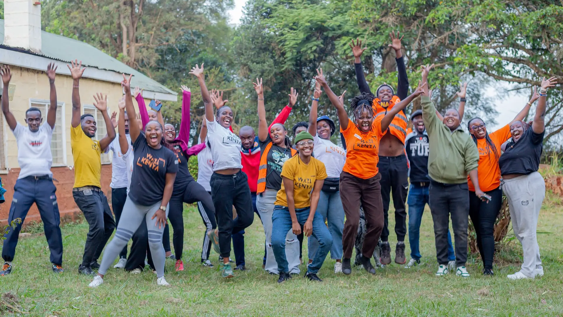Wazi volunteers leap together with arms raised on a green lawn.