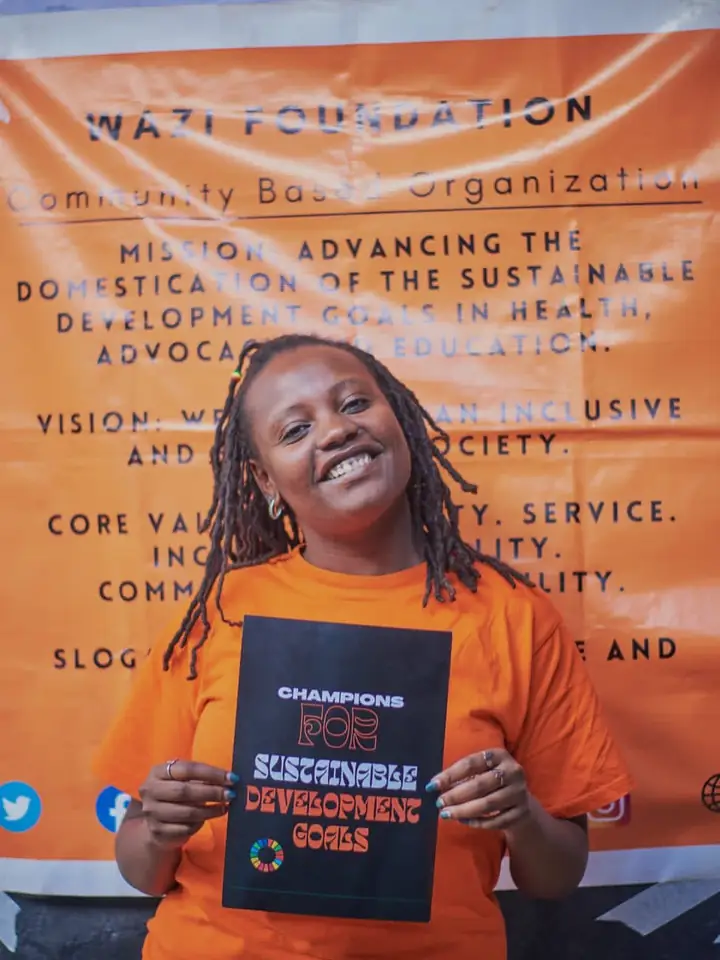 A laughing Wazi volunteer holds an SDG book in front of the orange organisation banner.